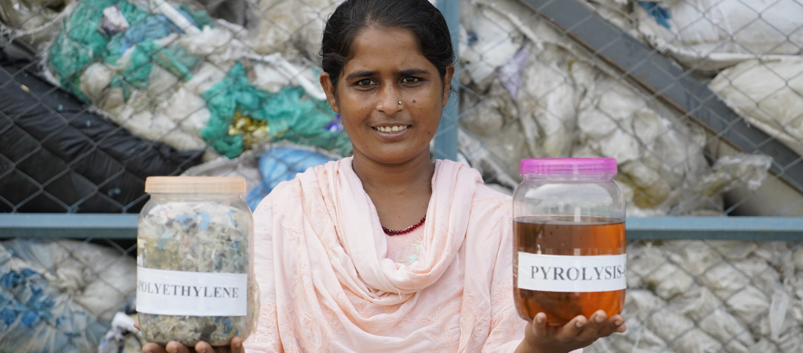 Nishi, a waste worker stands in front of a pile of plastic waste, holding a jar labeled "POLYETHYLENE" in one hand and a jar labeled "PYROLYSIS OIL" in the other.