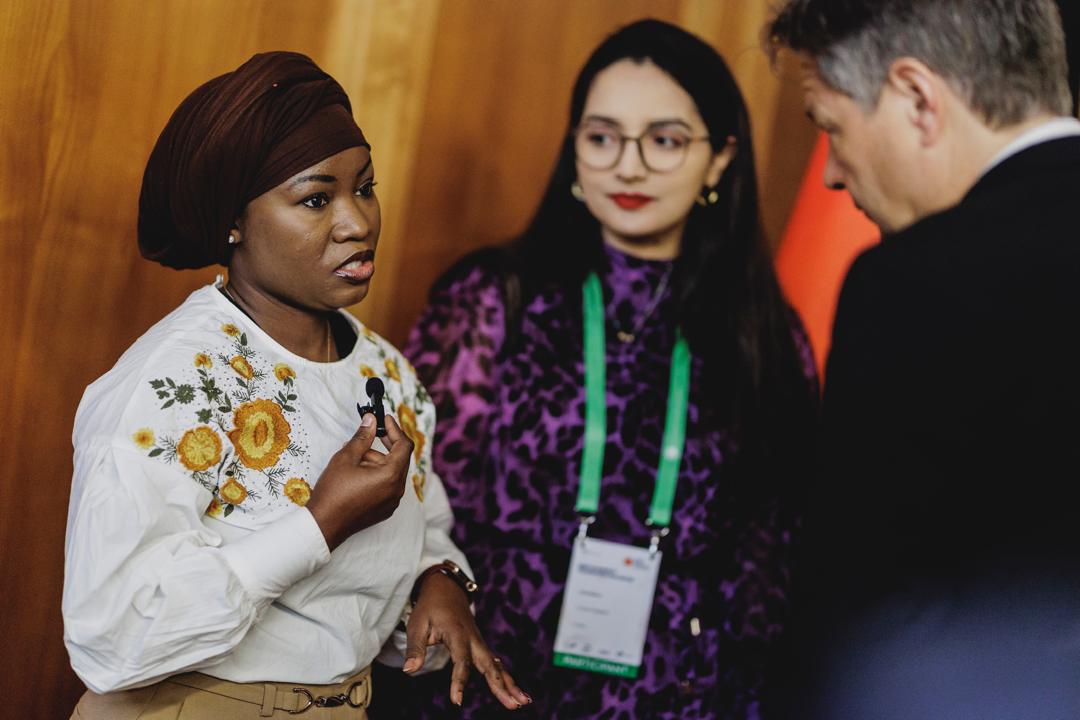 Three individuals engaged in conversation. A woman in a floral-embroidered white top and headscarf is speaking, while another woman in a purple outfit with glasses listens attentively. The man faces them.