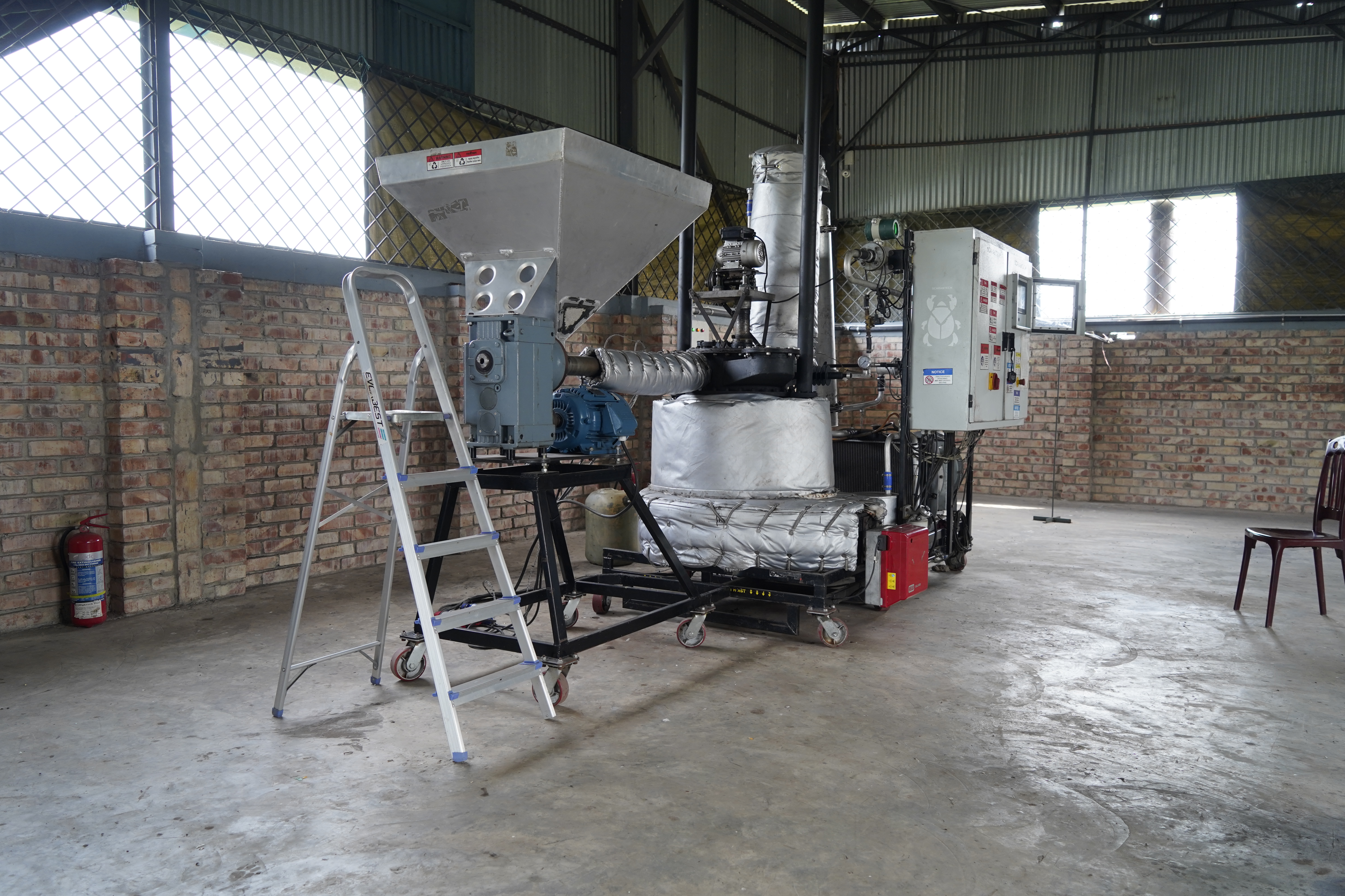 Pyrolysis machine - Industrial machinery with a metal hopper, control panel, and insulated pipes is set up inside a warehouse with brick walls and a concrete floor; a step ladder stands beside it.