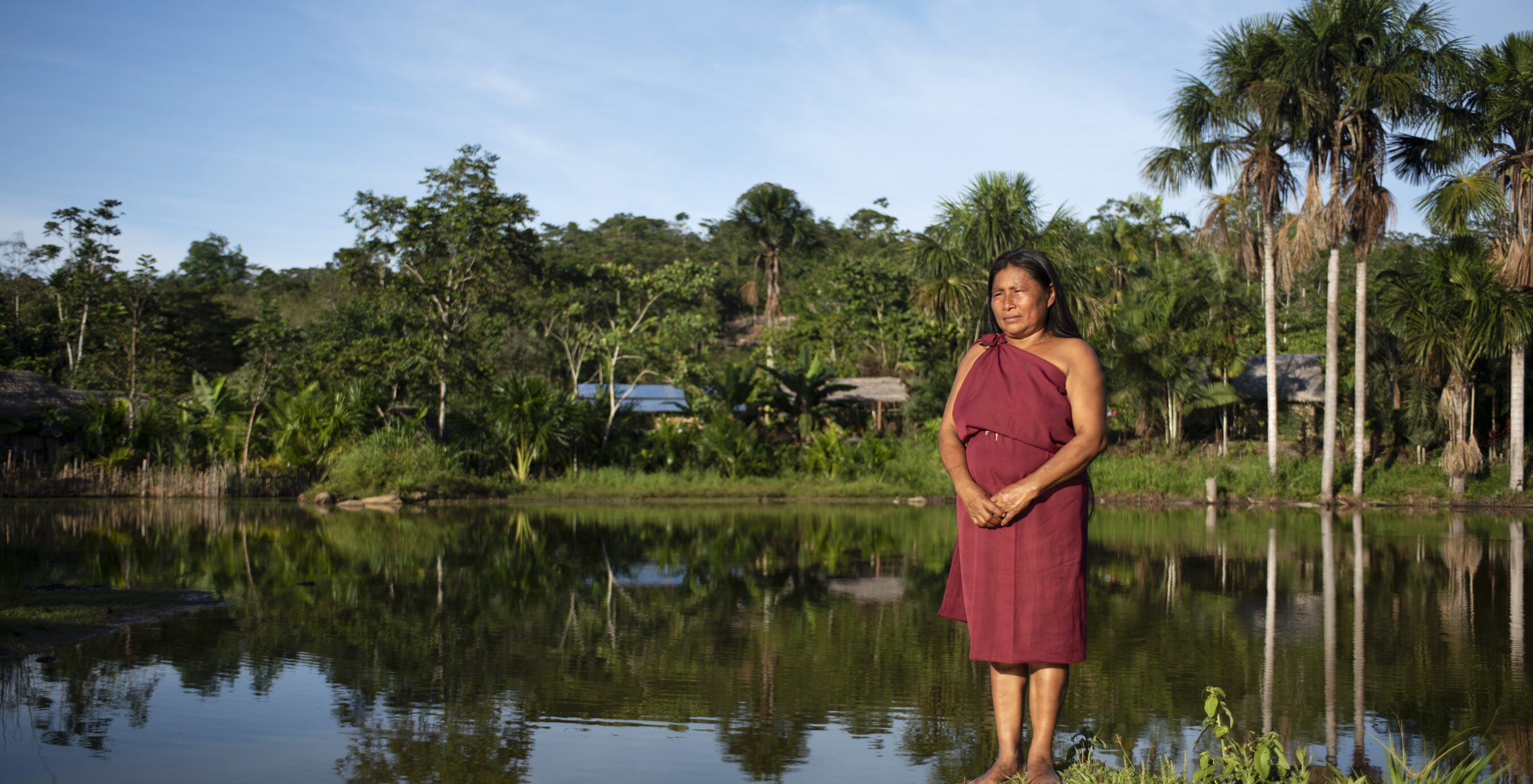 A woman in a maroon dress stands by a calm lake, surrounded by lush greenery and palm trees under a clear sky.
