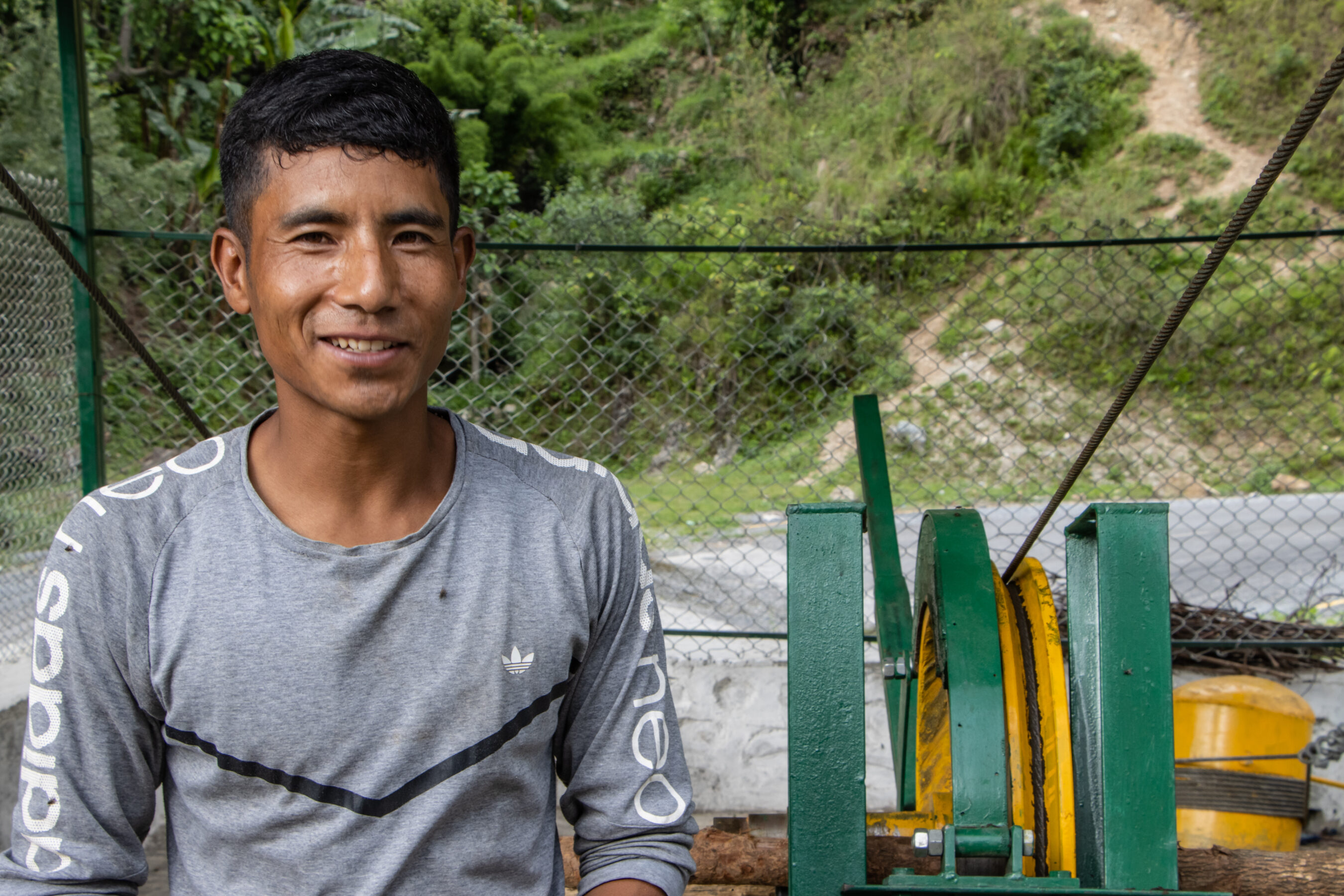 A man in a gray Adidas shirt smiles while standing outdoors near a green and yellow mechanical device, with a background of foliage and a fence.