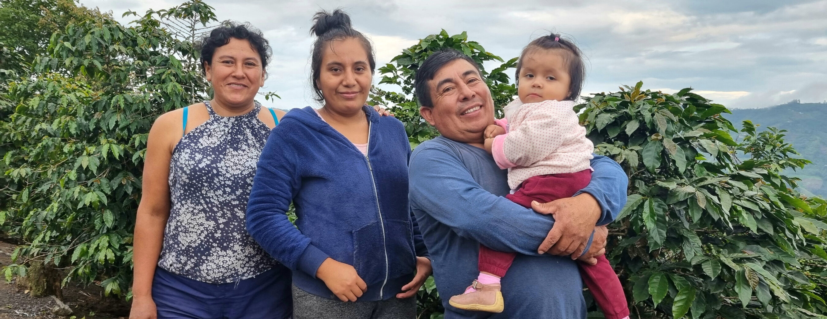A group of four people standing outdoors in front of greenery, including two women and a man holding a toddler.
