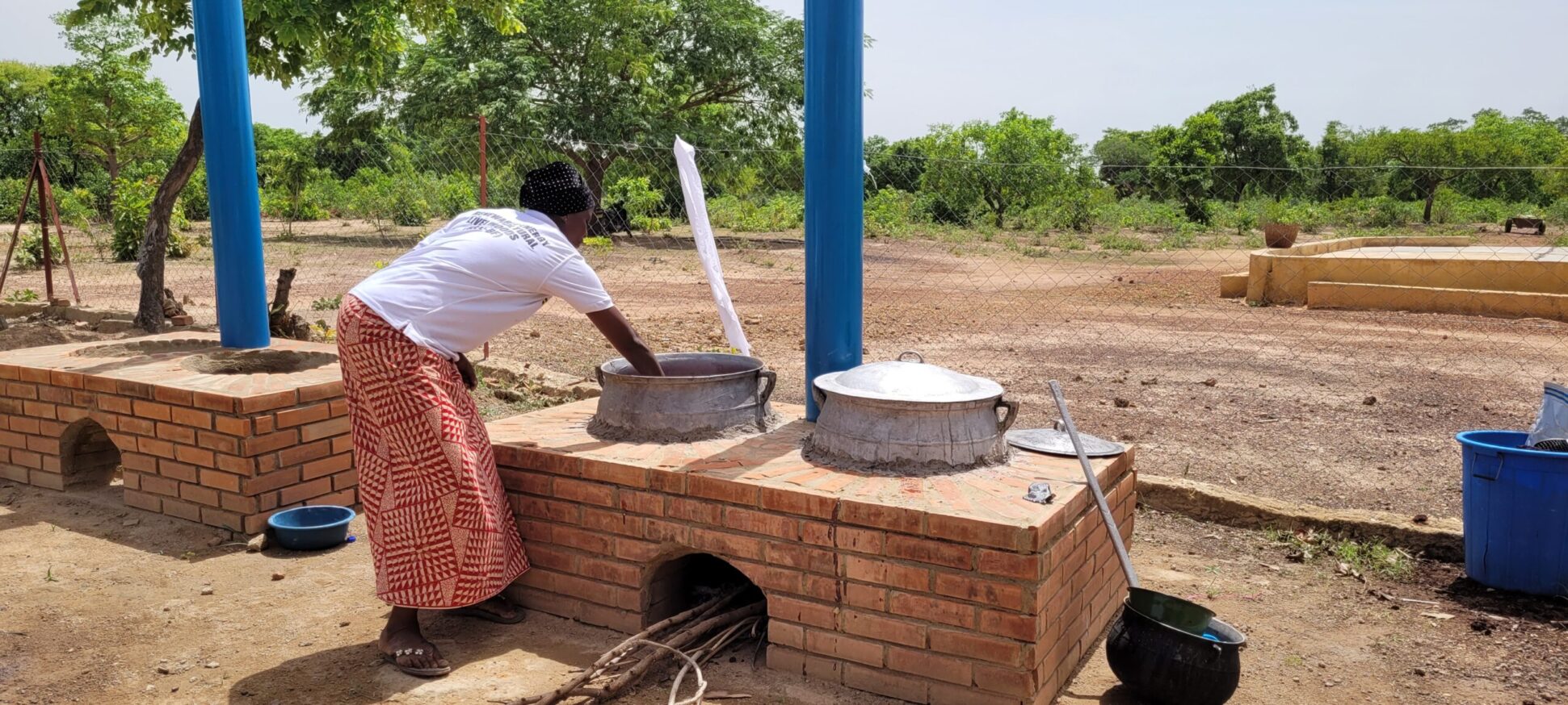 A person cooking outdoors on a brick stove with pots, surrounded by trees and open land.