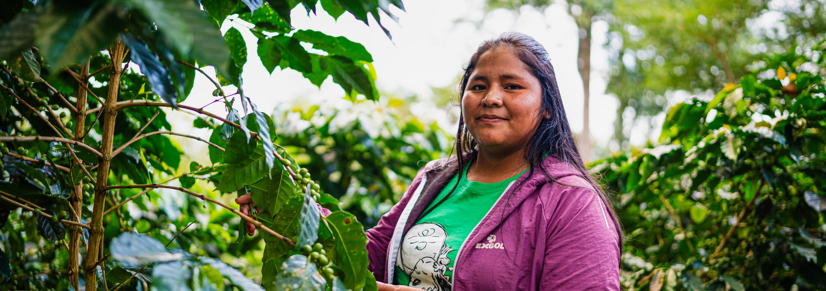 A woman in a purple jacket stands among green plants, smiling at the camera.