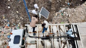 Two workers install equipment on a rooftop among scattered debris. A solar panel and other technical devices are visible. The ground around appears cluttered with trash.