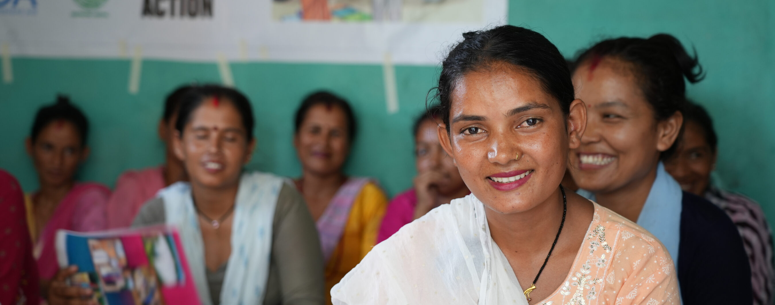 Puspa Sunars with a group of women, some smiling, sit together in a room with teal walls and a banner partially visible in the background.