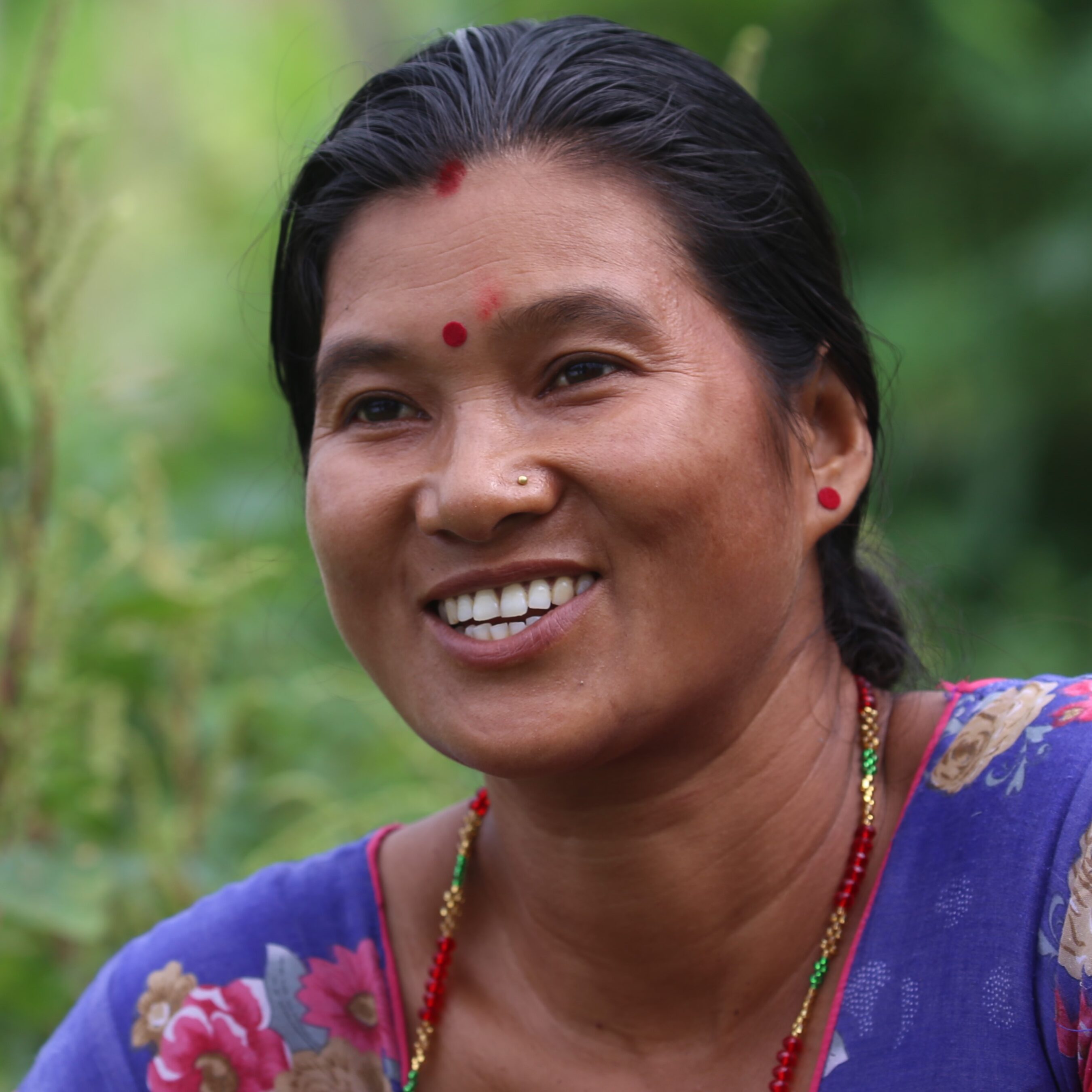 A woman wearing a colorful floral blouse and red jewelry smiles in a green outdoor setting.
