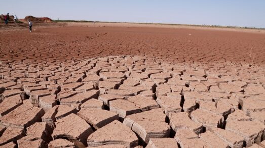 Cracked and dry earth under a clear sky, indicating severe drought conditions. A few people stand in the distant background.