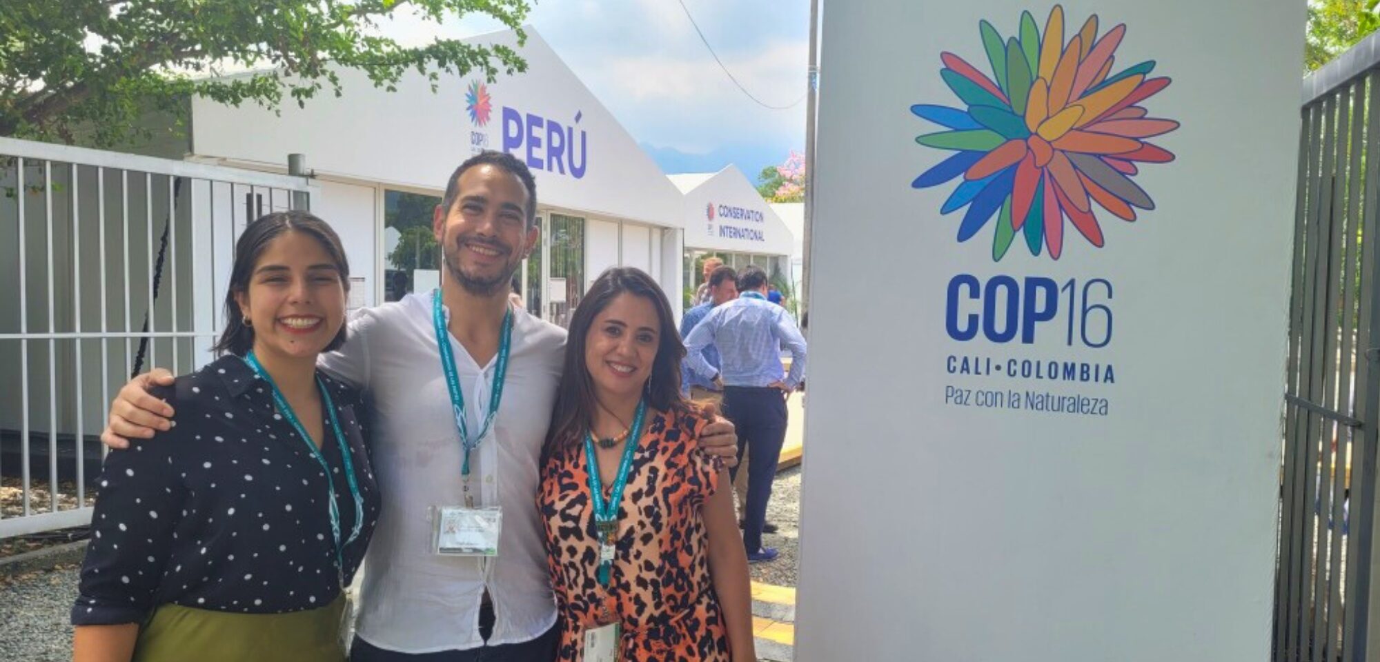 Three people standing and smiling in front of a sign for COP16 in Cali, Colombia, with "Paz con la Naturaleza" and colorful logo.