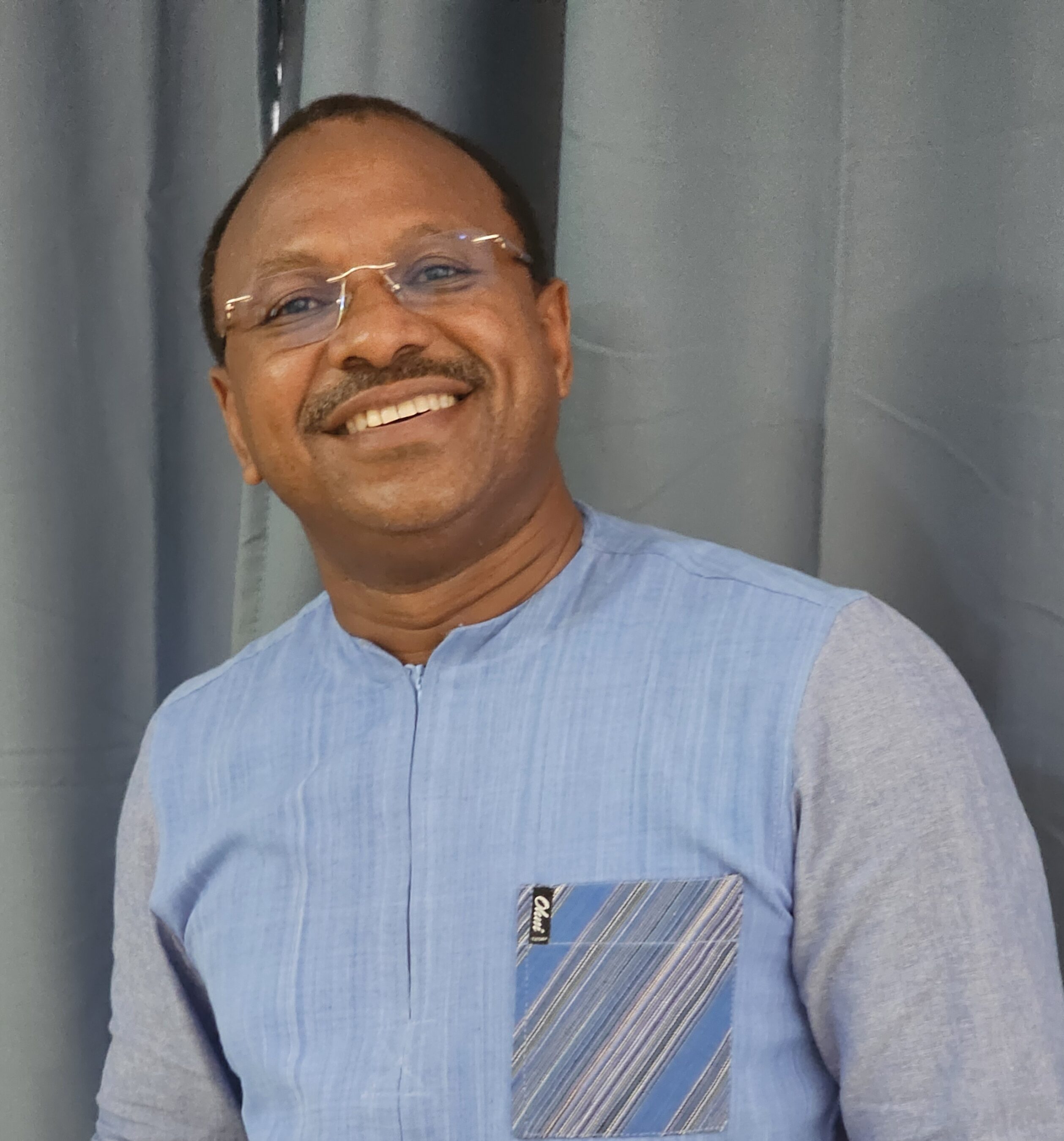 Smiling man wearing glasses and a light blue shirt with a striped pocket standing in front of a gray curtain.