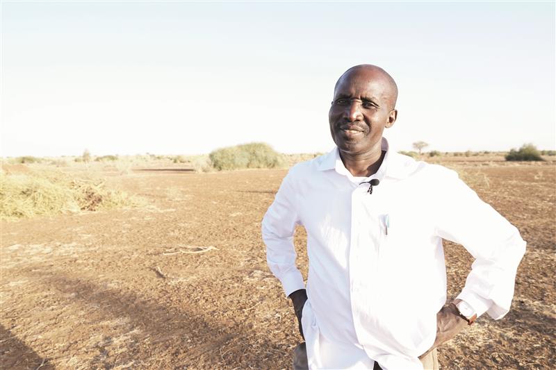 A man in a white shirt stands with hands on hips in a dry, open landscape under a clear sky.
