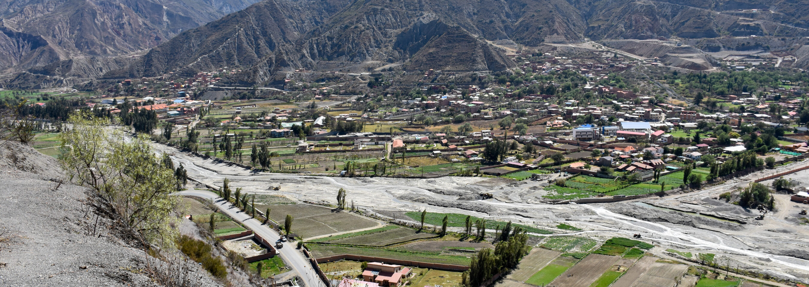 Aerial view of a mountainous landscape with a village, riverbed, and surrounding greenery under a clear sky.