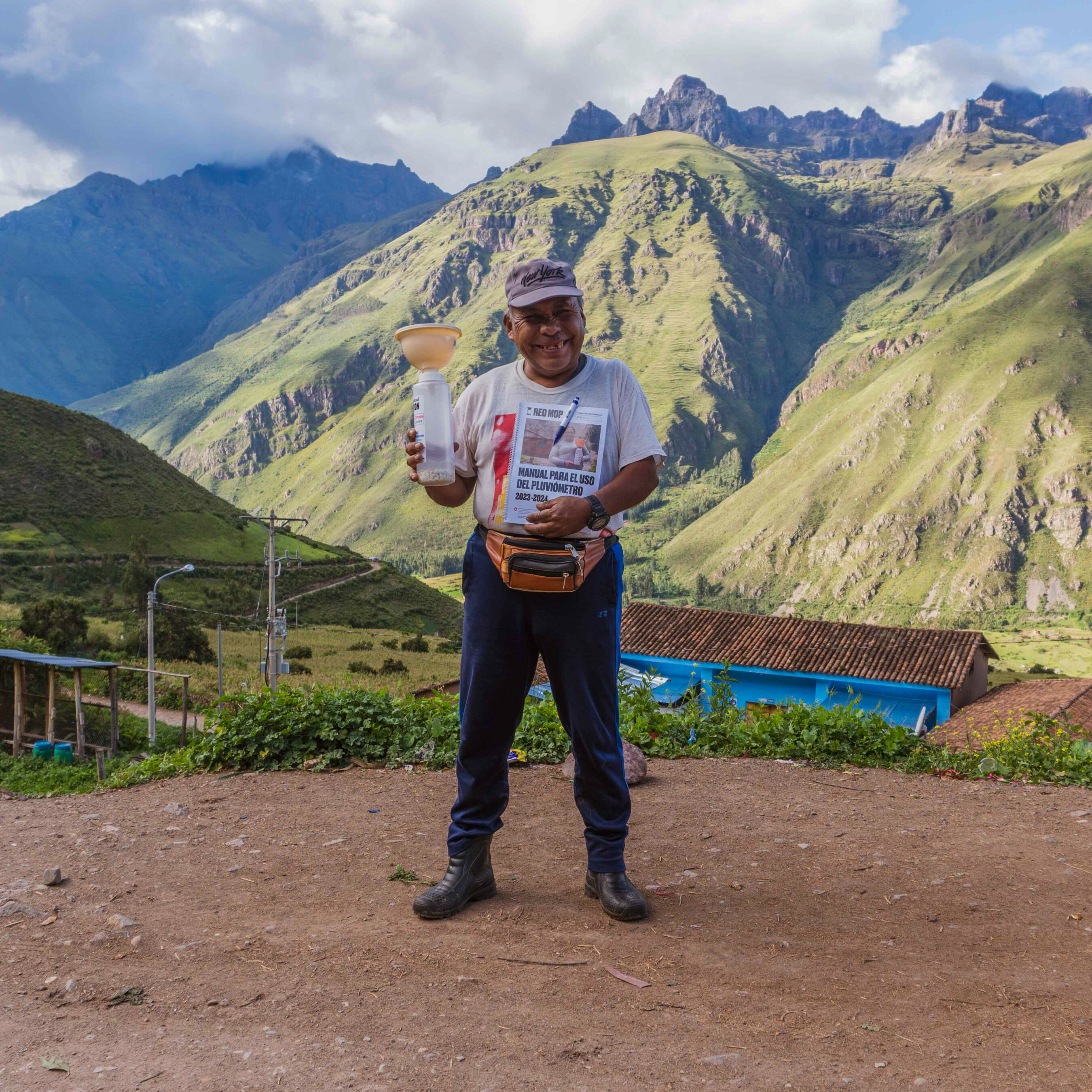 A man wearing a cap and t-shirt stands on a dirt path in front of a mountain landscape, holding a large light bulb.