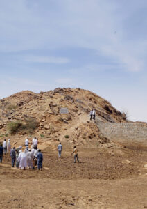 A group of people stand near a rocky hill with a stone wall under a blue sky. Some climb the hill. Sparse vegetation is visible.