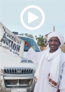 Man in traditional attire stands next to a white Hyundai SUV, holding a flag with 