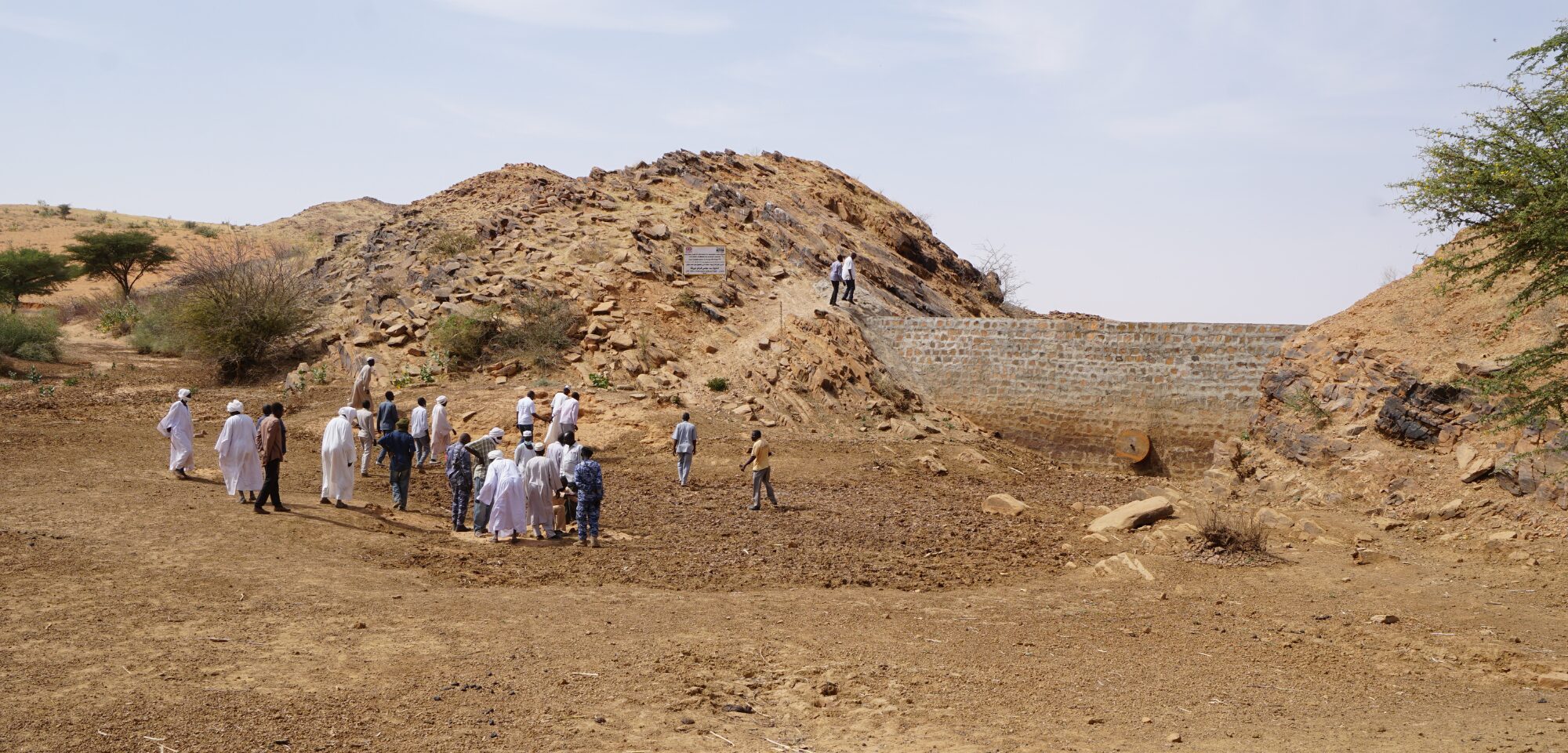 A group of people stands near a dry dam and rocky mound in an arid landscape under a clear sky.