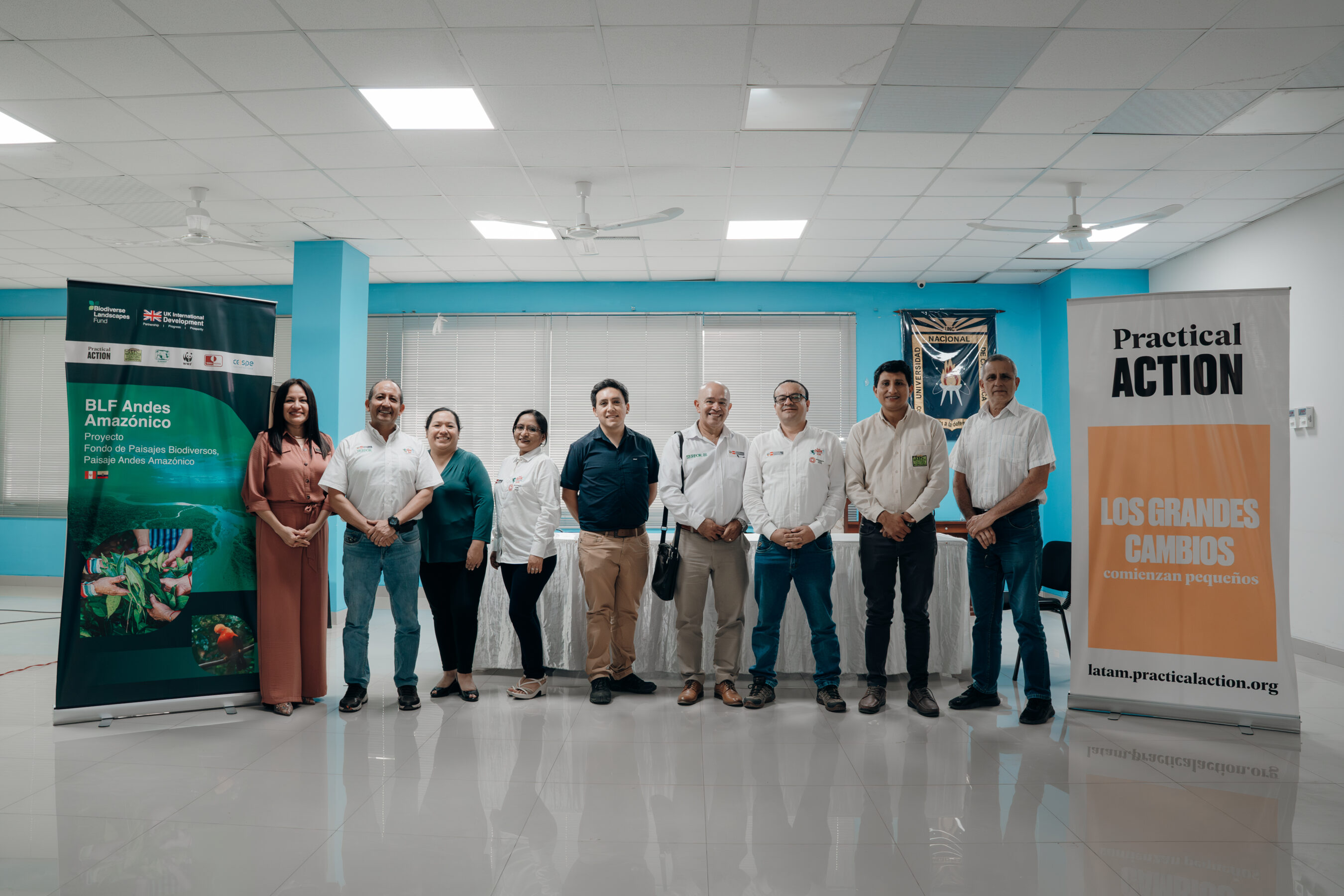 A group of ten people stands indoors, posing for a photo between two informational banners in a brightly lit room with tiled floors and blue walls.