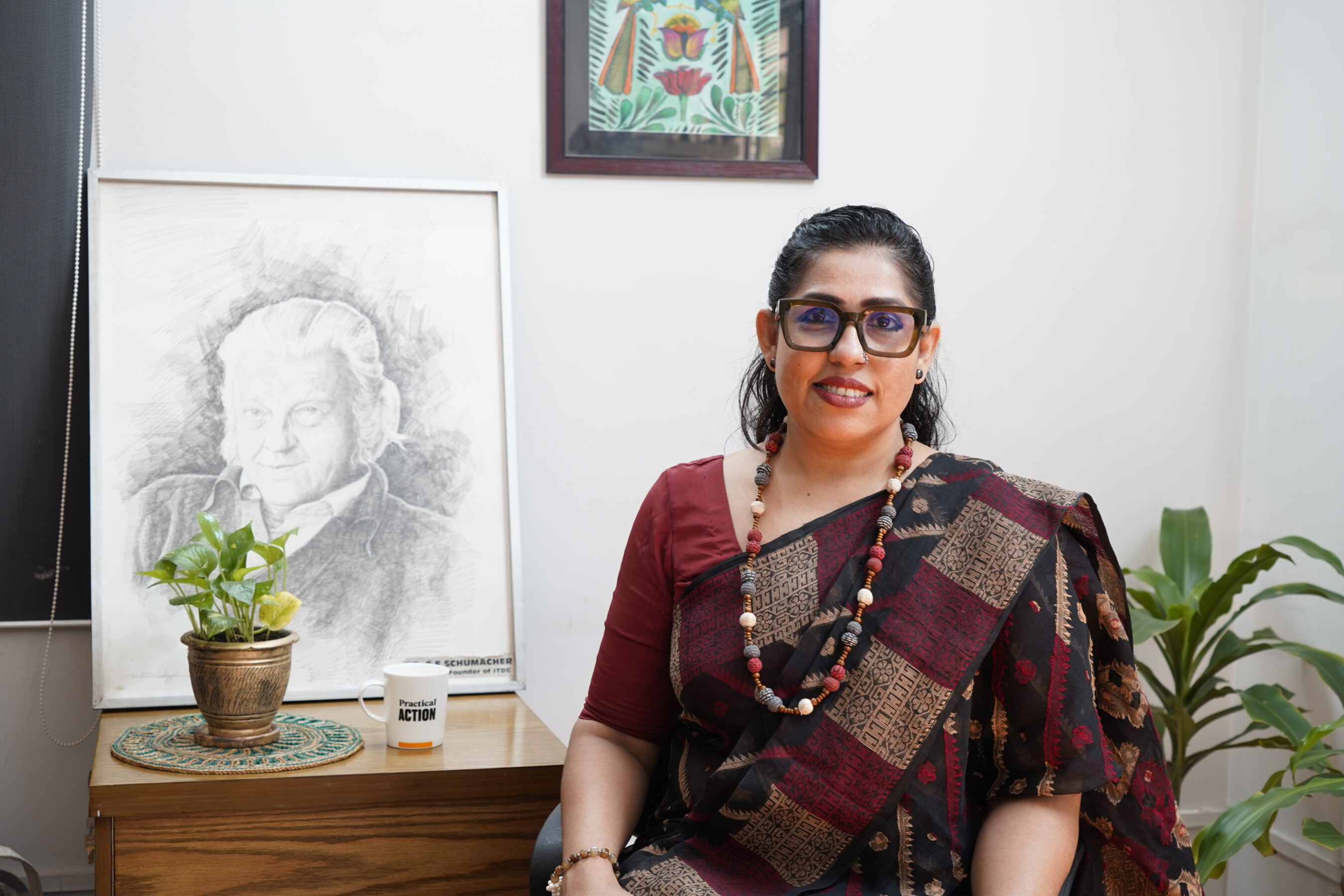 A person in a patterned sari sits beside a framed sketch, with a plant and a mug on the table. A colorful artwork hangs on the wall behind.