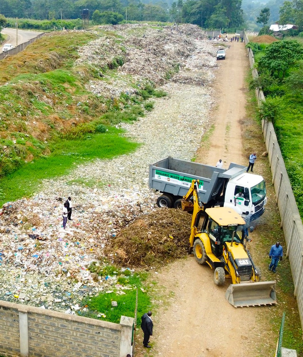 Aerial view of a landfill with a bulldozer and a truck working. People stand nearby. The area is bordered by a wall and greenery is visible in the background.