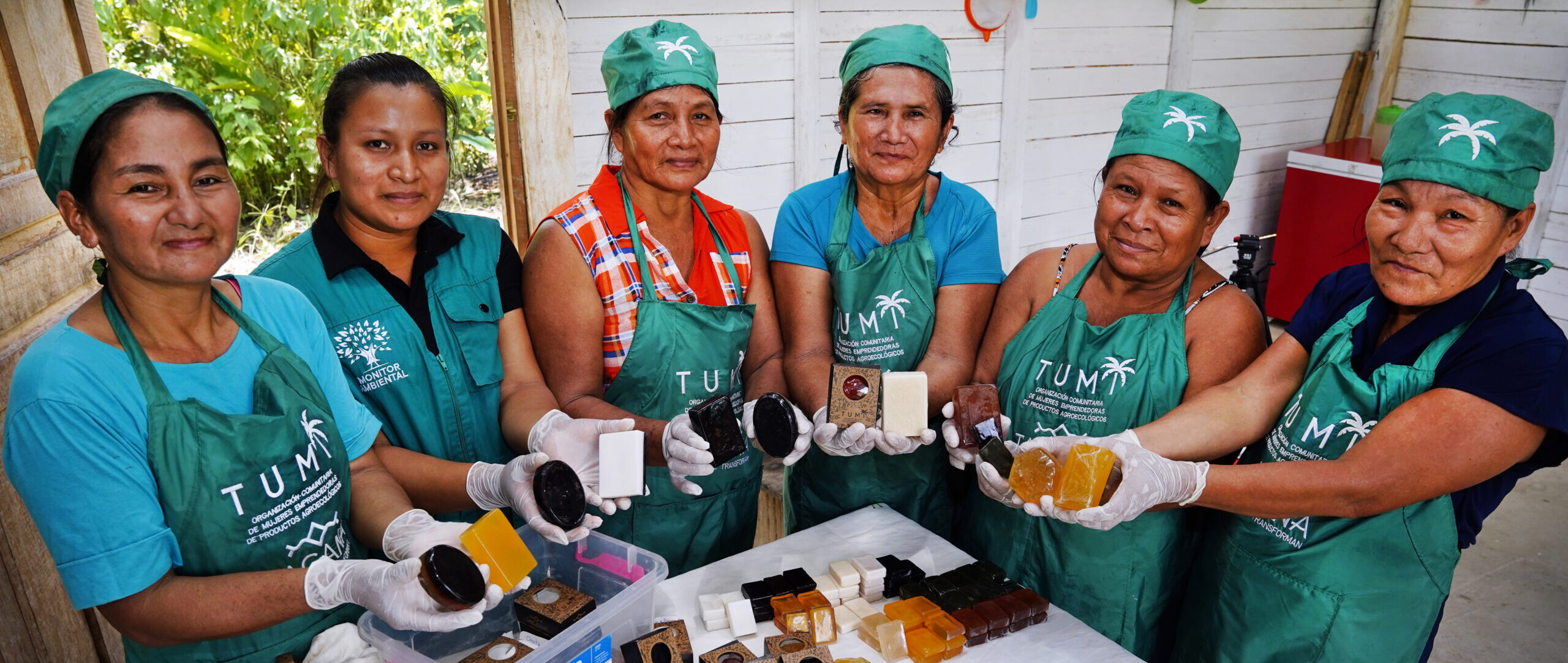 A group of women in aprons proudly display handmade soap products in a workshop setting. The table in front of them holds various soap bars.