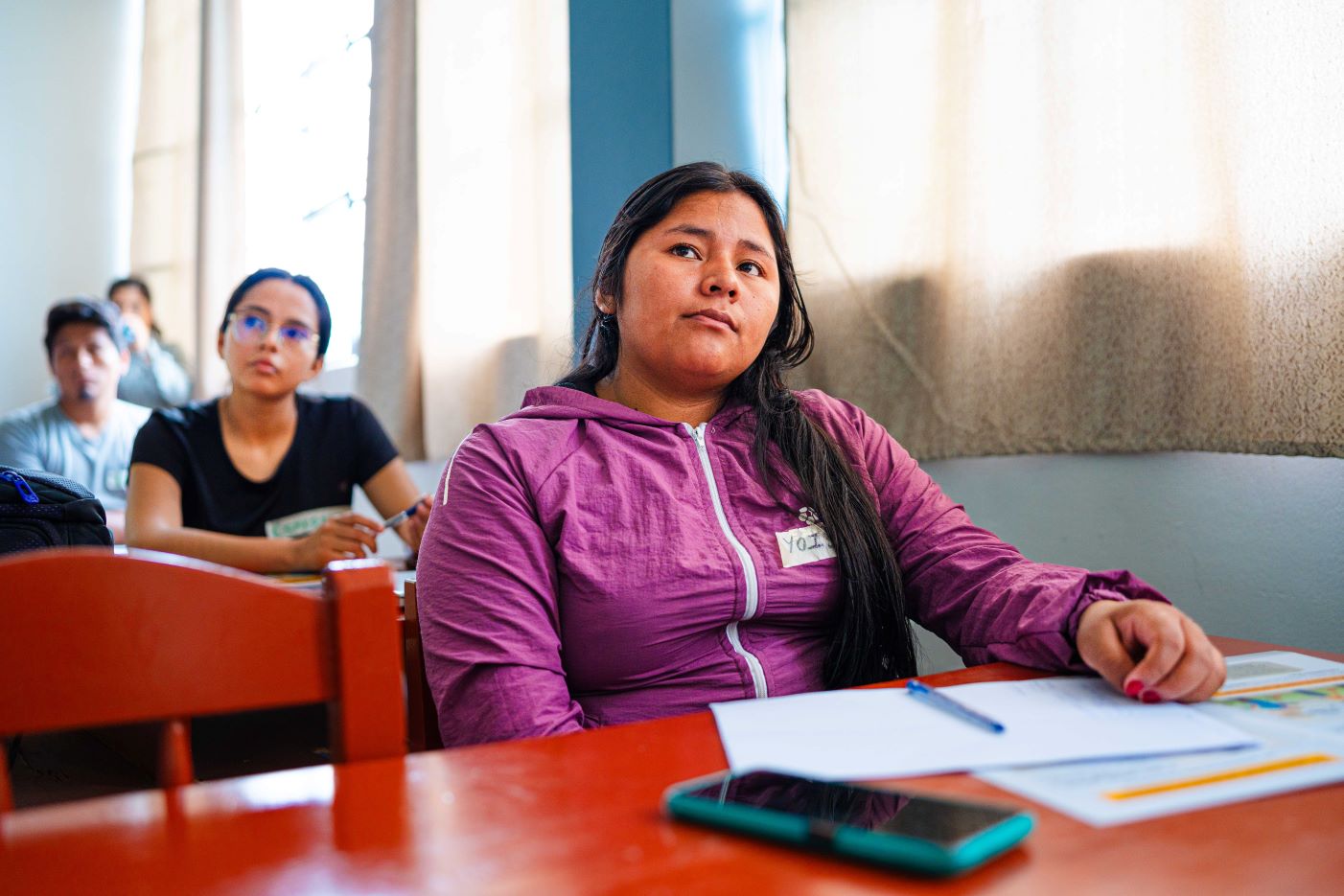 Person in a purple jacket sits in a classroom, looking forward attentively. A notebook, pen, and smartphone are on the desk. Other students are visible in the background.