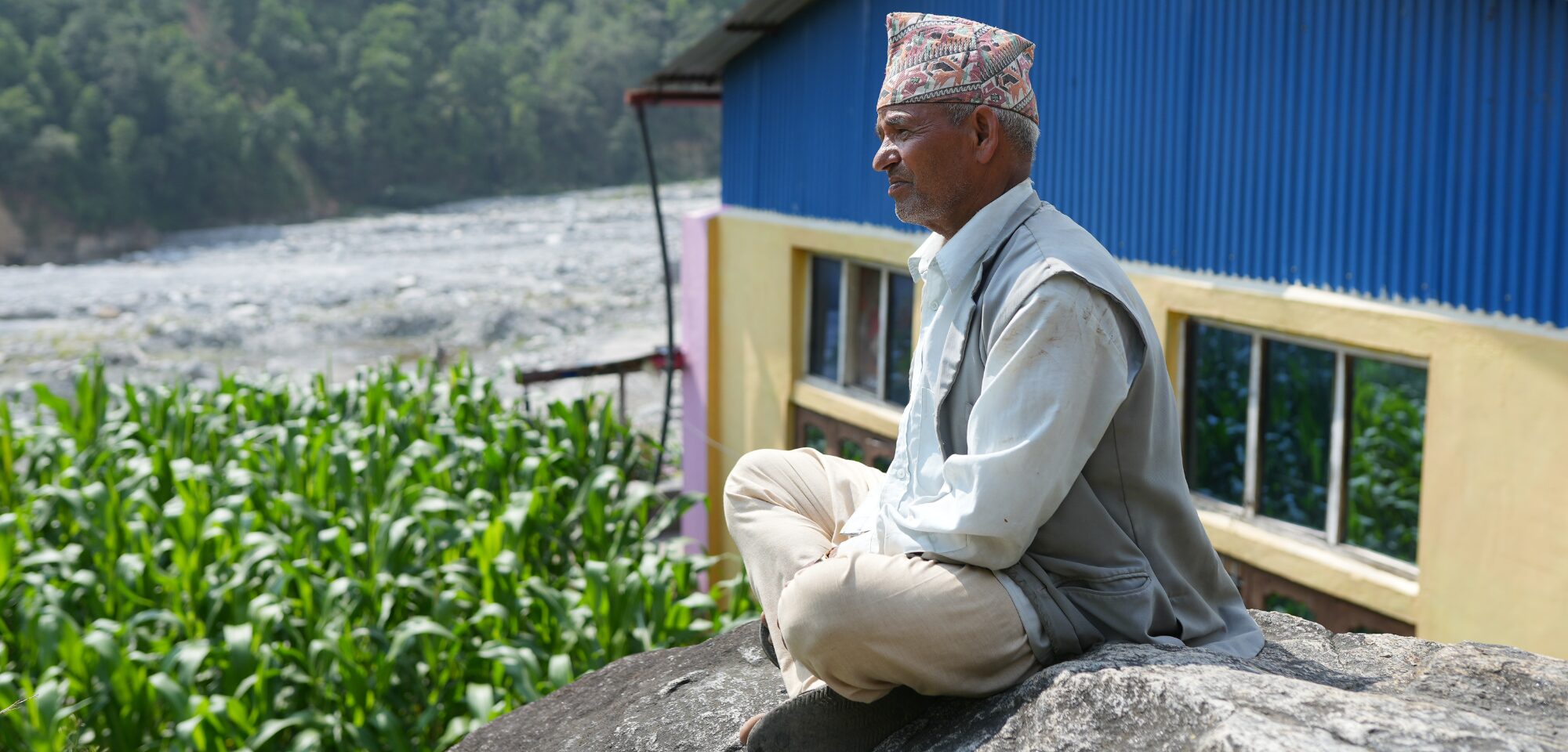 An elderly man wearing a traditional hat sits on a rock, overlooking a lush green field and a river next to a blue-roofed building.