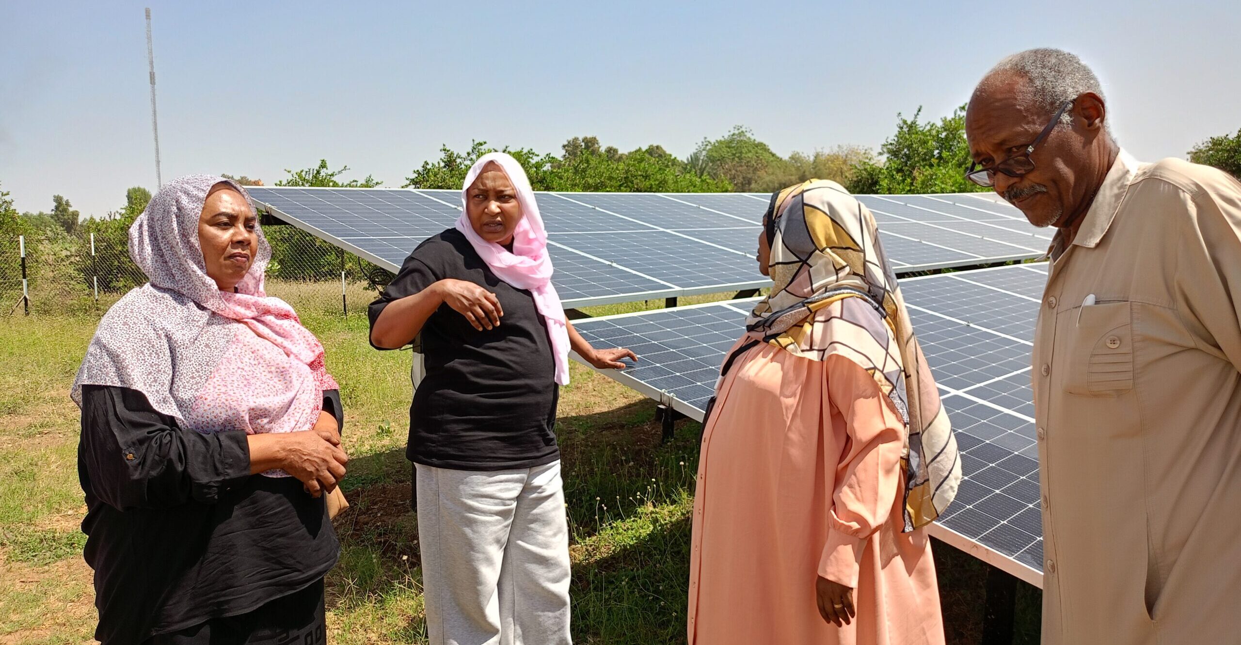 Four people stand in front of solar panels, engaged in conversation. They are outdoors on a sunny day with greenery in the background.