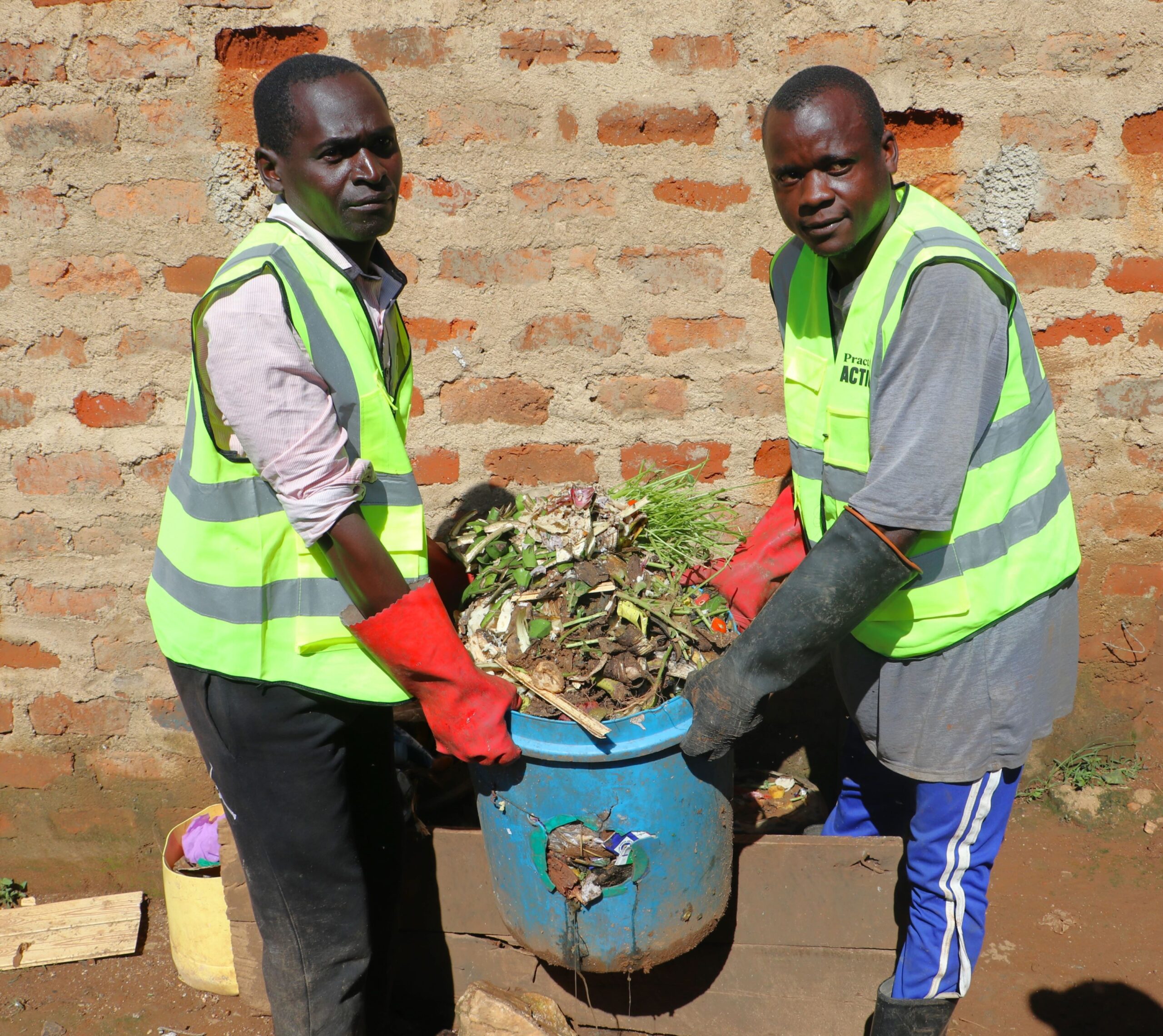Two men wearing reflective vests and gloves carry a blue container filled with compost material in front of a brick wall.
