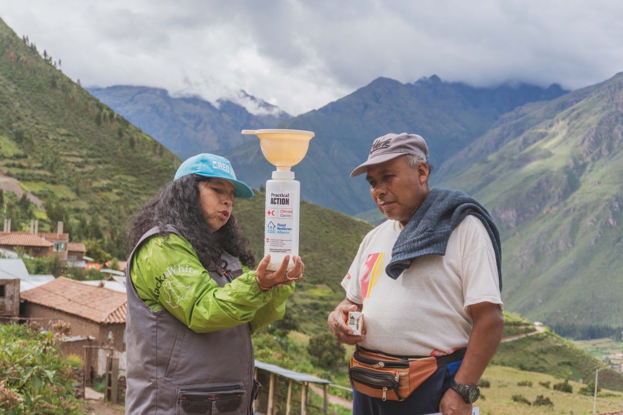 Two people stand outdoors in a mountainous area, examining a plastic bottle with a filter or funnel attached. They appear engaged in discussion or demonstration.