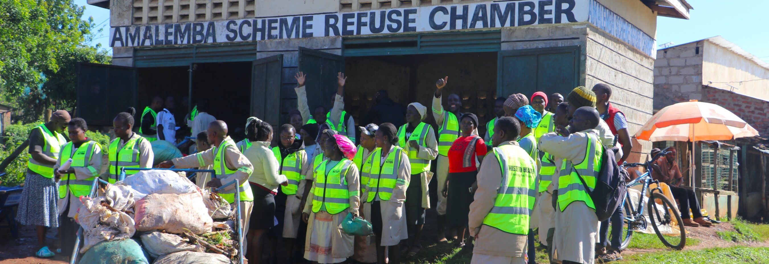 A group of people in high-visibility vests gather near a refuse chamber labeled "Amalembo Scheme Refuse Chamber." Trash is visible in the foreground.