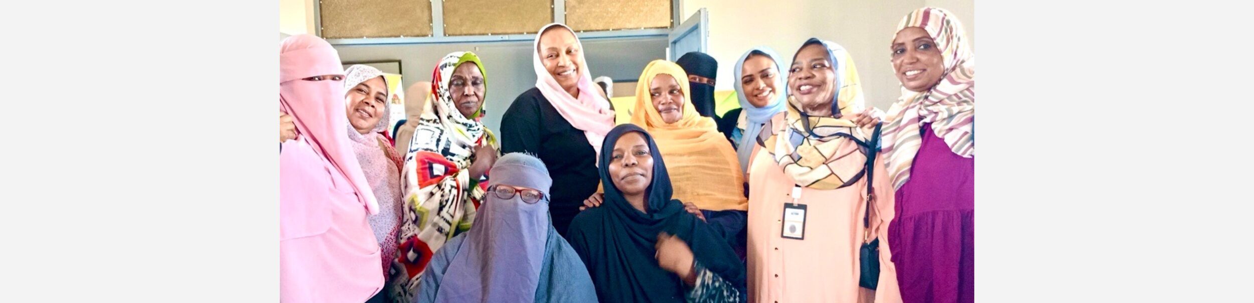A group of women, some wearing hijabs, are posed together in a room. They are standing and sitting closely, smiling, with a mix of light and dark clothing.