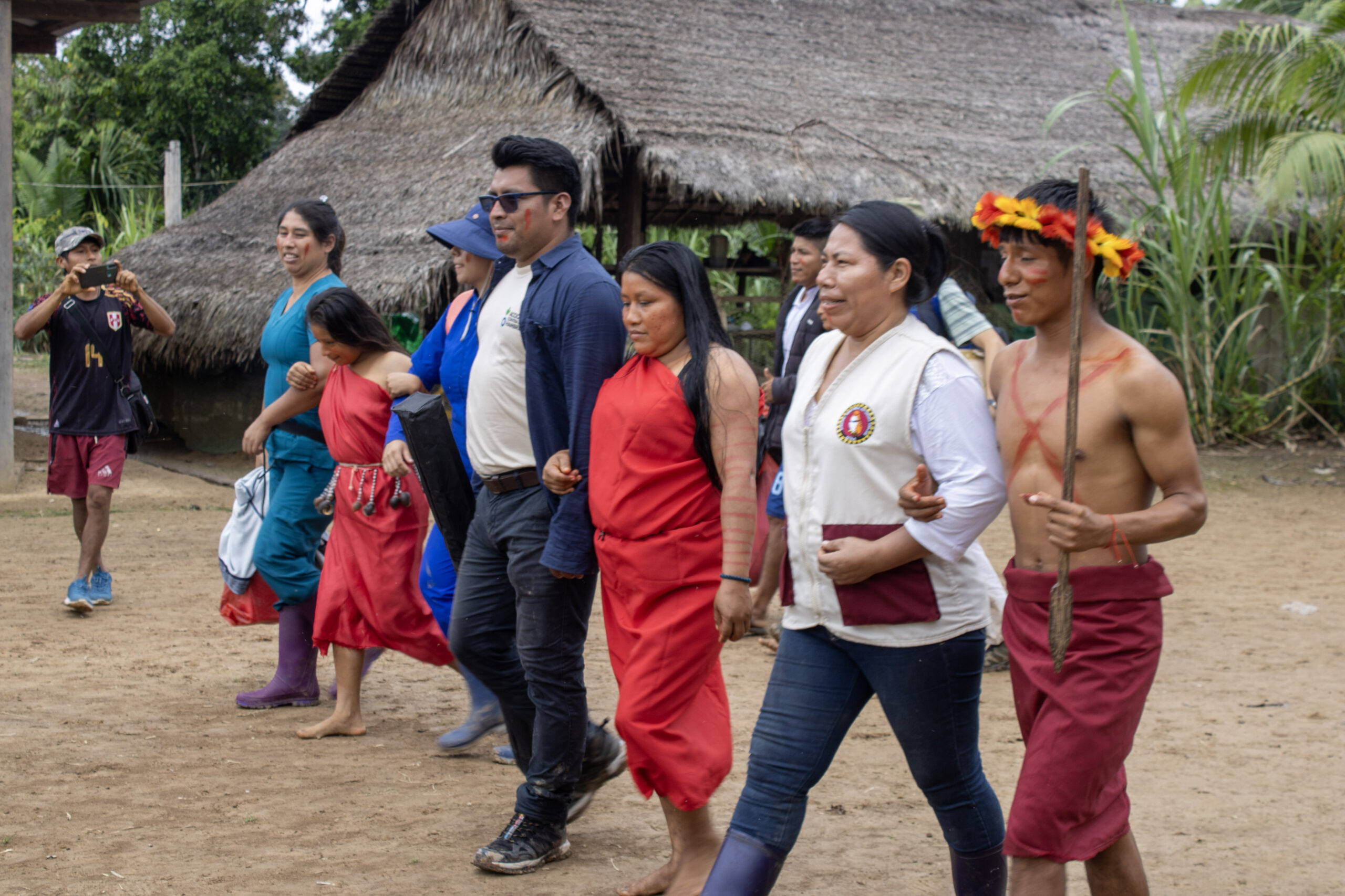 A group of people, including individuals in traditional attire, walk together outdoors near a thatched-roof building, with one person in the background taking a photo.
