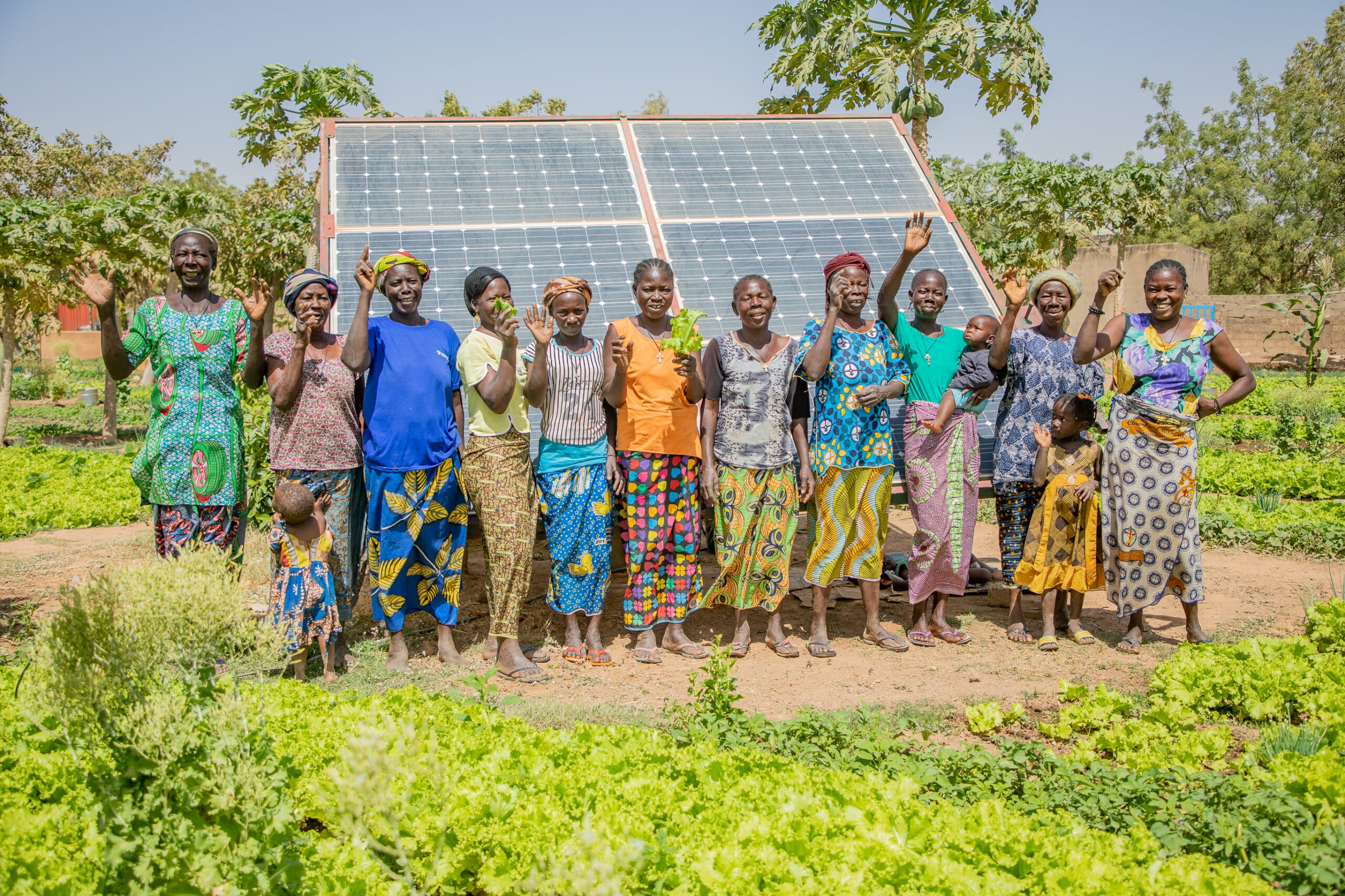 A group of people stand in front of a solar panel in a lush garden, smiling and waving.