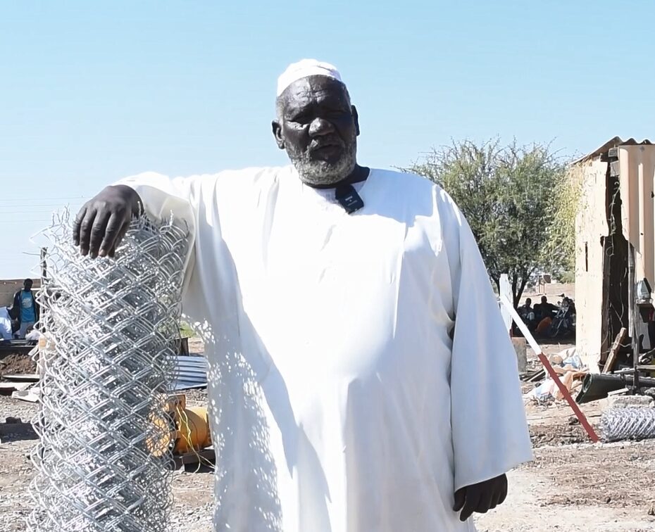 Man in a white robe and cap stands beside a coiled metal fence outdoors, with a clear sky and scattered structures in the background.