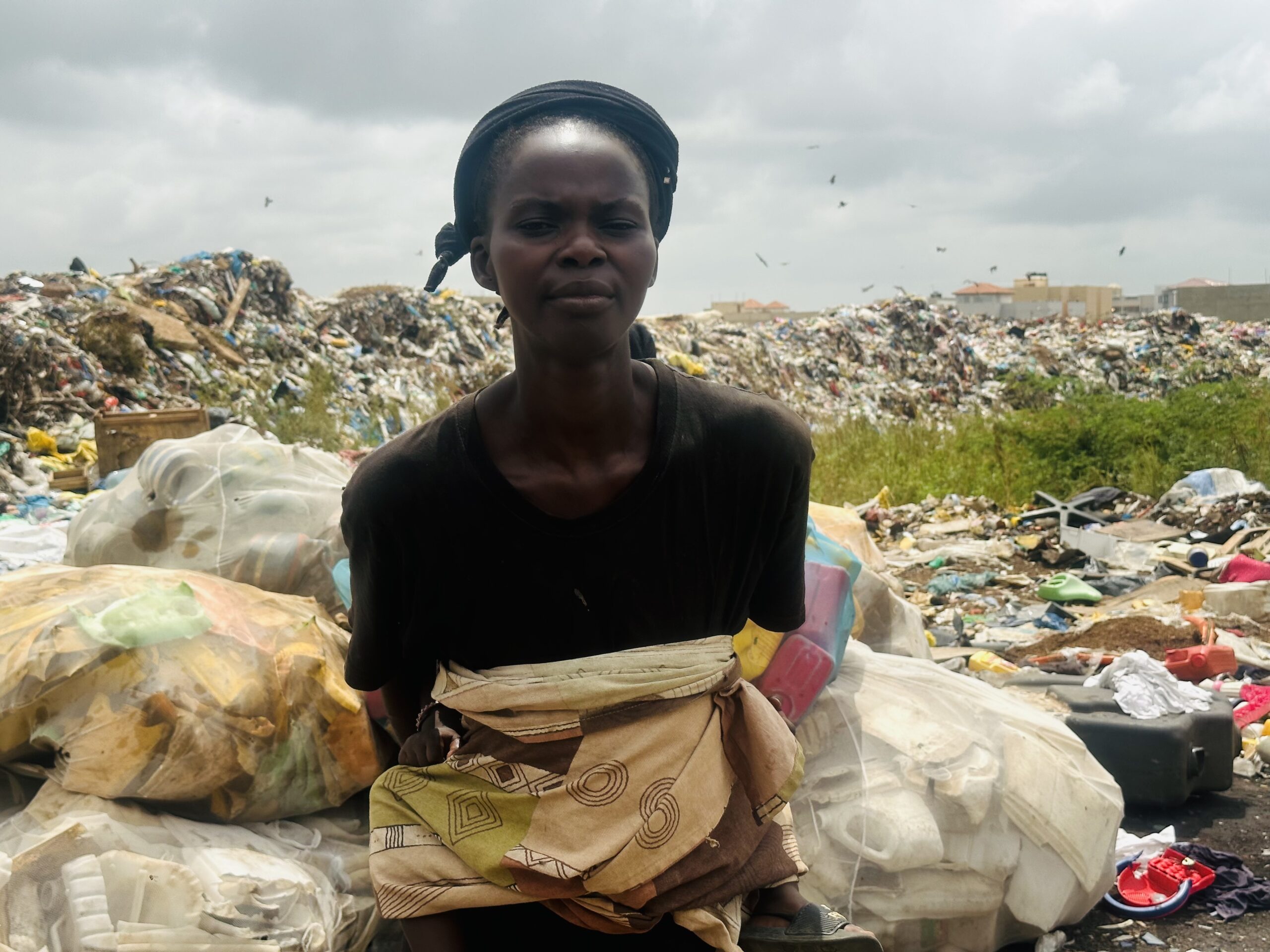 Person standing in front of a large pile of trash, wearing a black shirt and patterned fabric tied around the waist, under overcast skies.