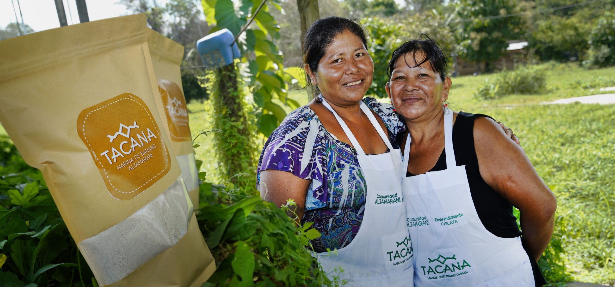 Two women wearing aprons stand outdoors holding a bag with the label 'TACANA.' They are surrounded by greenery, smiling at the camera.
