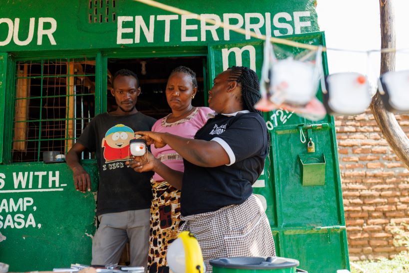 Three people stand in front of a green shop. One person is holding and explaining a solar lamp, while the others listen. A line with several solar lamps hangs in the foreground.