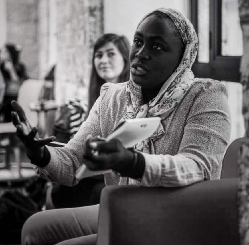 A woman wearing a headscarf speaks while gesturing with her hands, holding a tablet. Another person sits in the background, listening. Black and white photo.