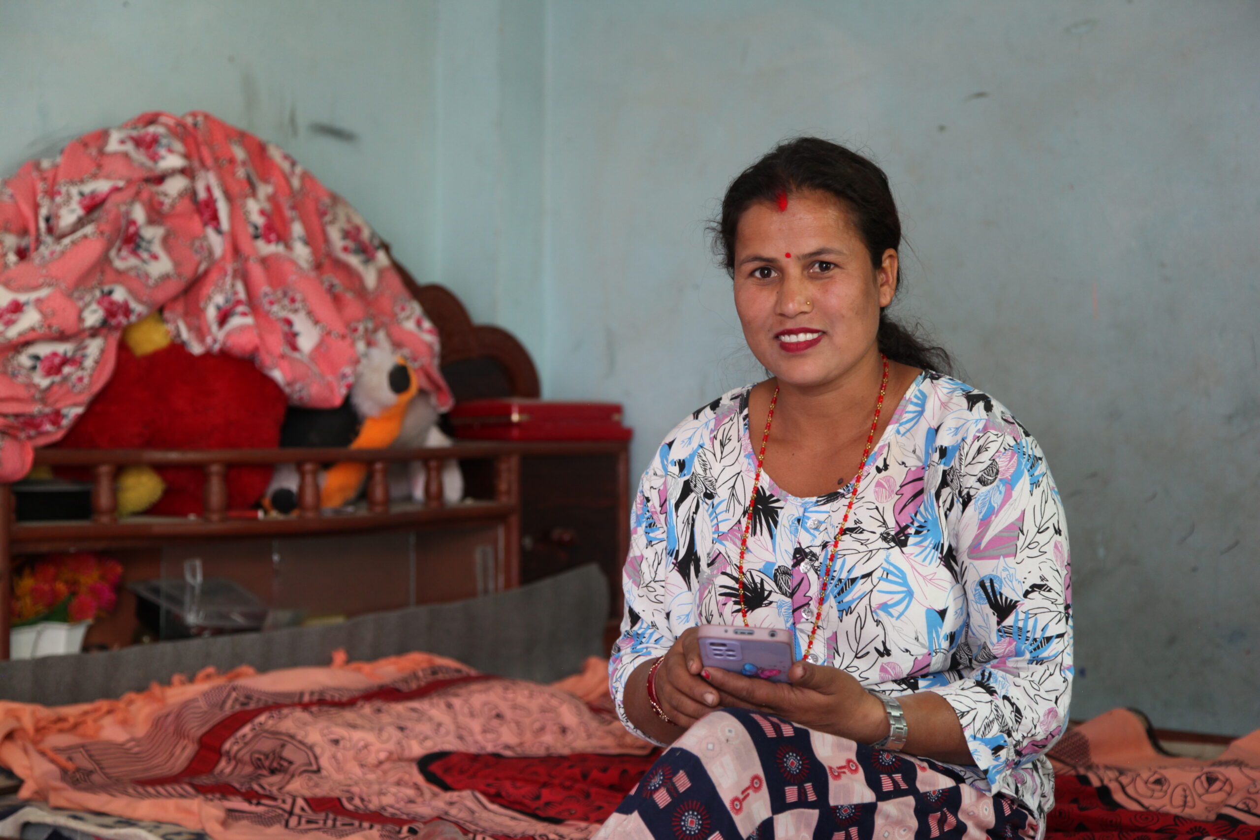 A woman in a floral dress sits on a bed, smiling and holding a smartphone. The room contains a bed with patterned bedding and stuffed toys on a shelf.