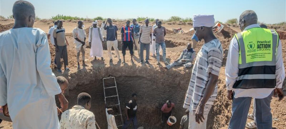 A group of people stand around a large pit in a dry, rural area, with some individuals wearing reflective vests, and a ladder is visible leading into the pit.