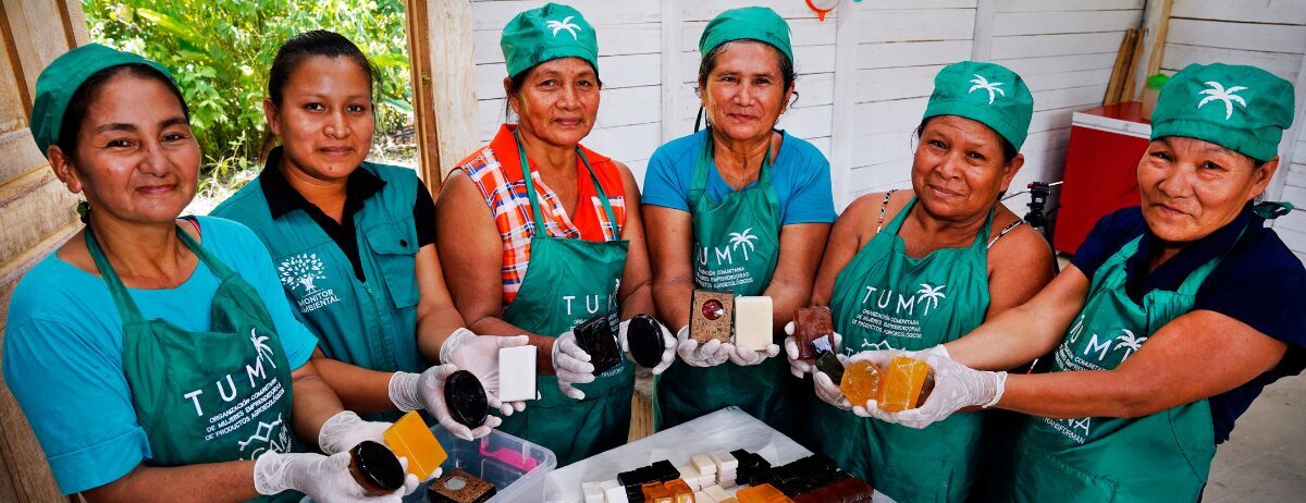 A group of six women in green aprons and hats display handmade soap bars in a room with white walls.