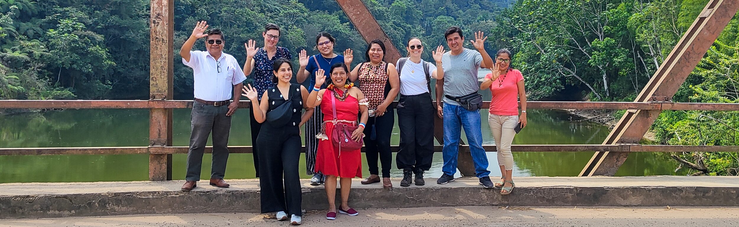 A group of nine people stands on a bridge, smiling and waving. Trees and a river are in the background.