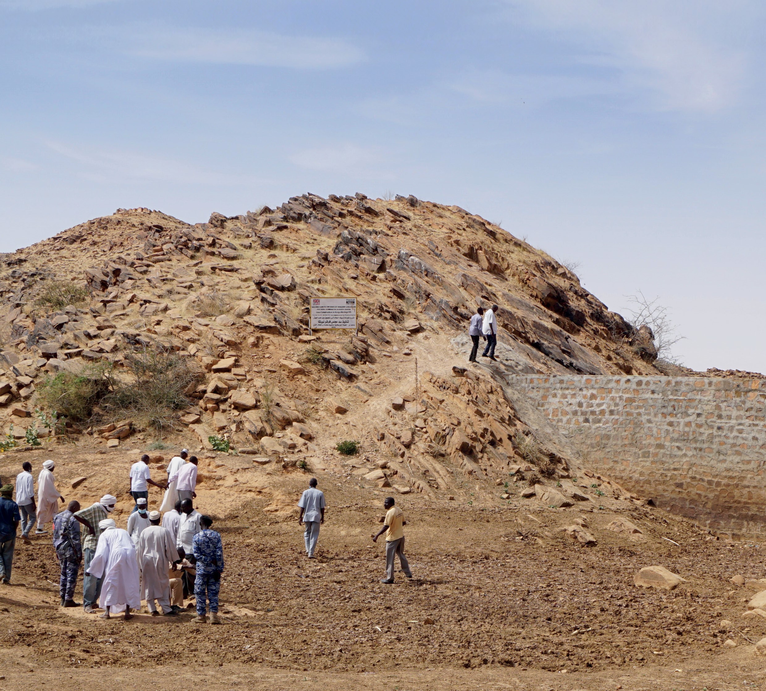 A group of people stands and watches as a few individuals walk up a rocky hill under a clear sky.
