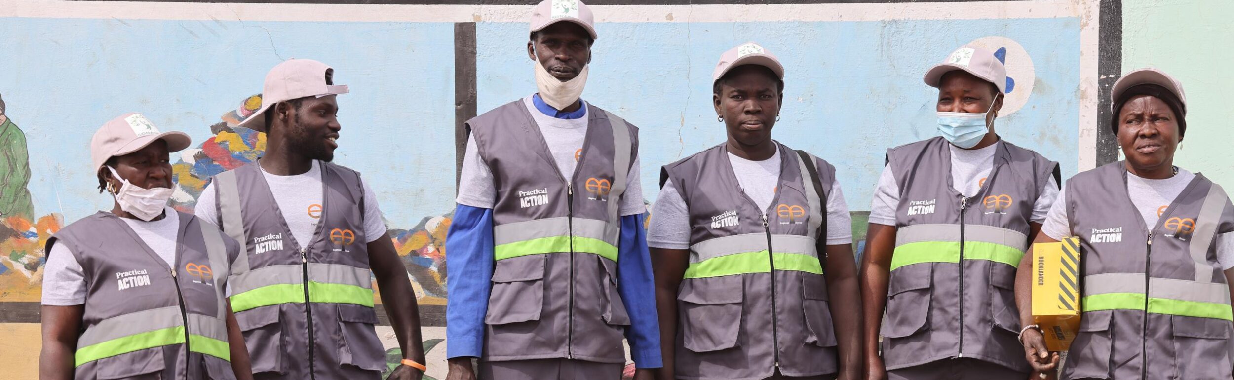 A group of six people in uniform with reflective vests and hats stand in front of a mural.