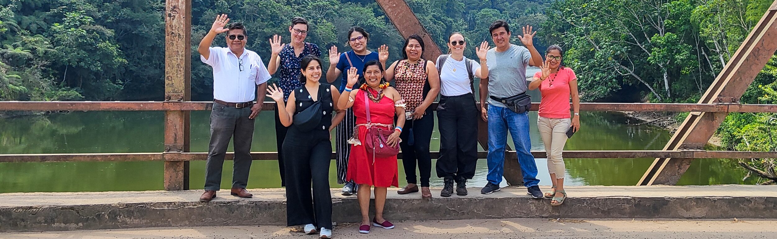 A group of people standing and waving on a bridge, with a lush green forest and river in the background.