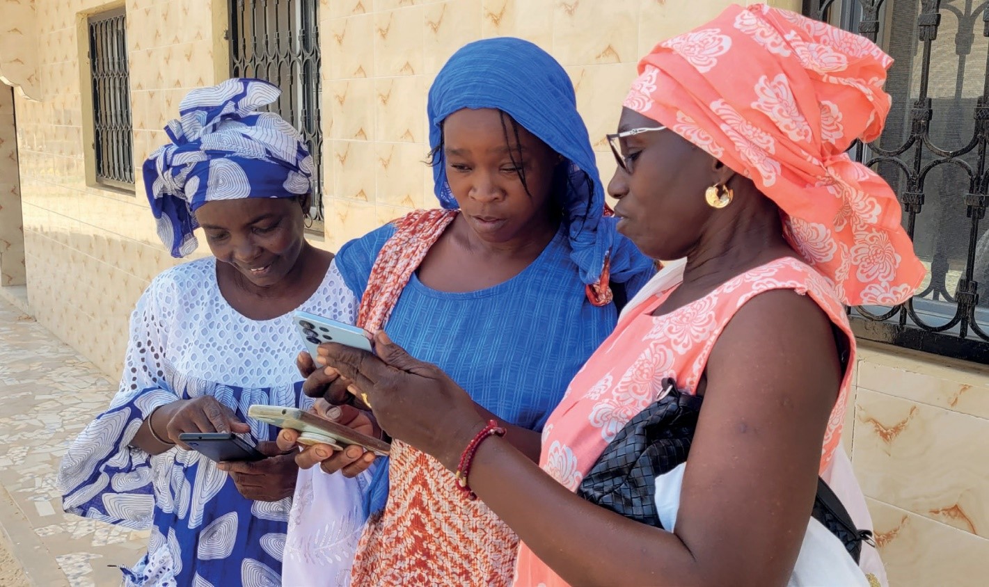 Three women in colorful traditional attire stand together outdoors, looking at their smartphones.
