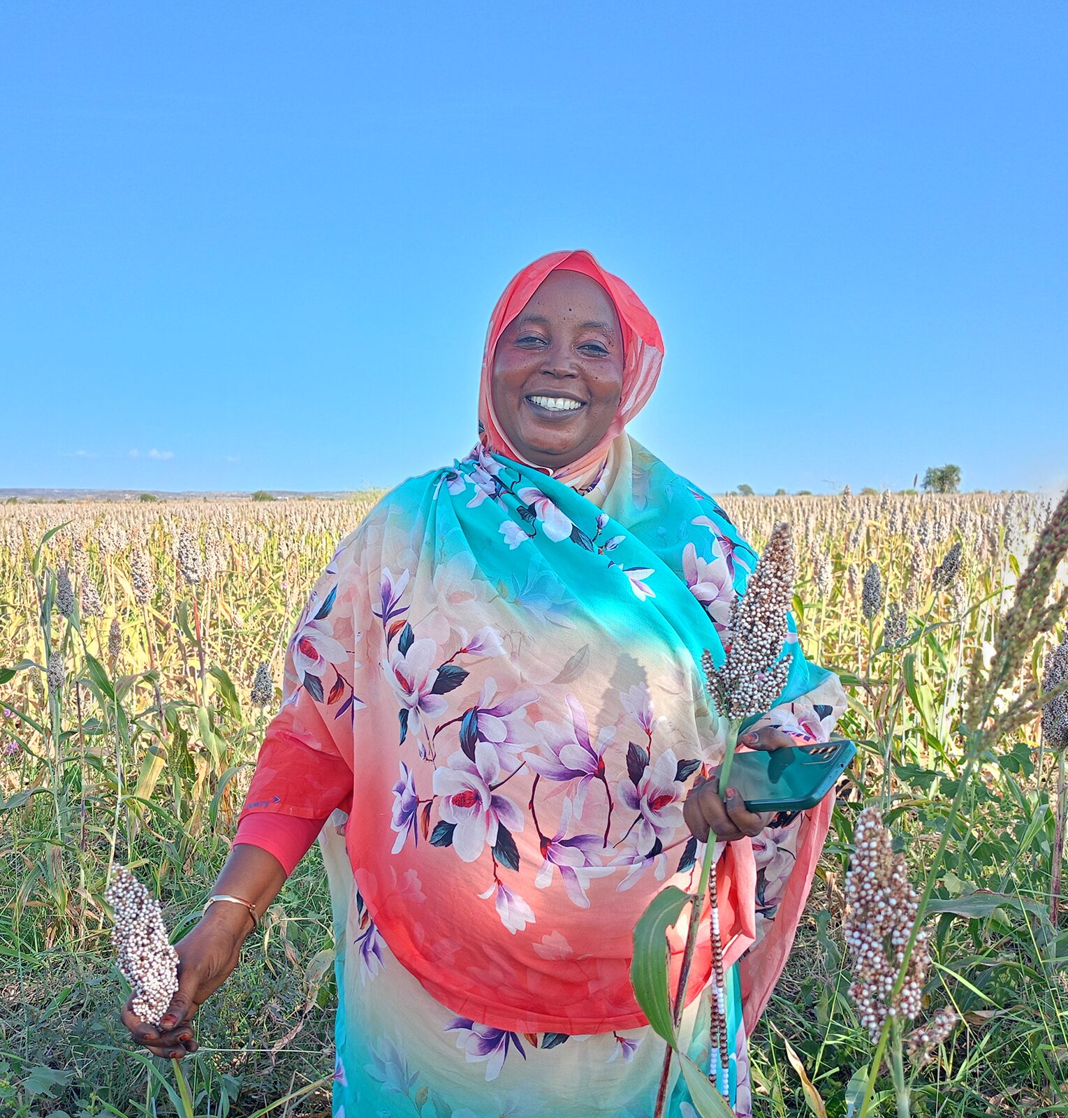 A woman in a colorful floral dress and headscarf stands smiling in a sorghum field, holding harvested sorghum plants under a clear blue sky.