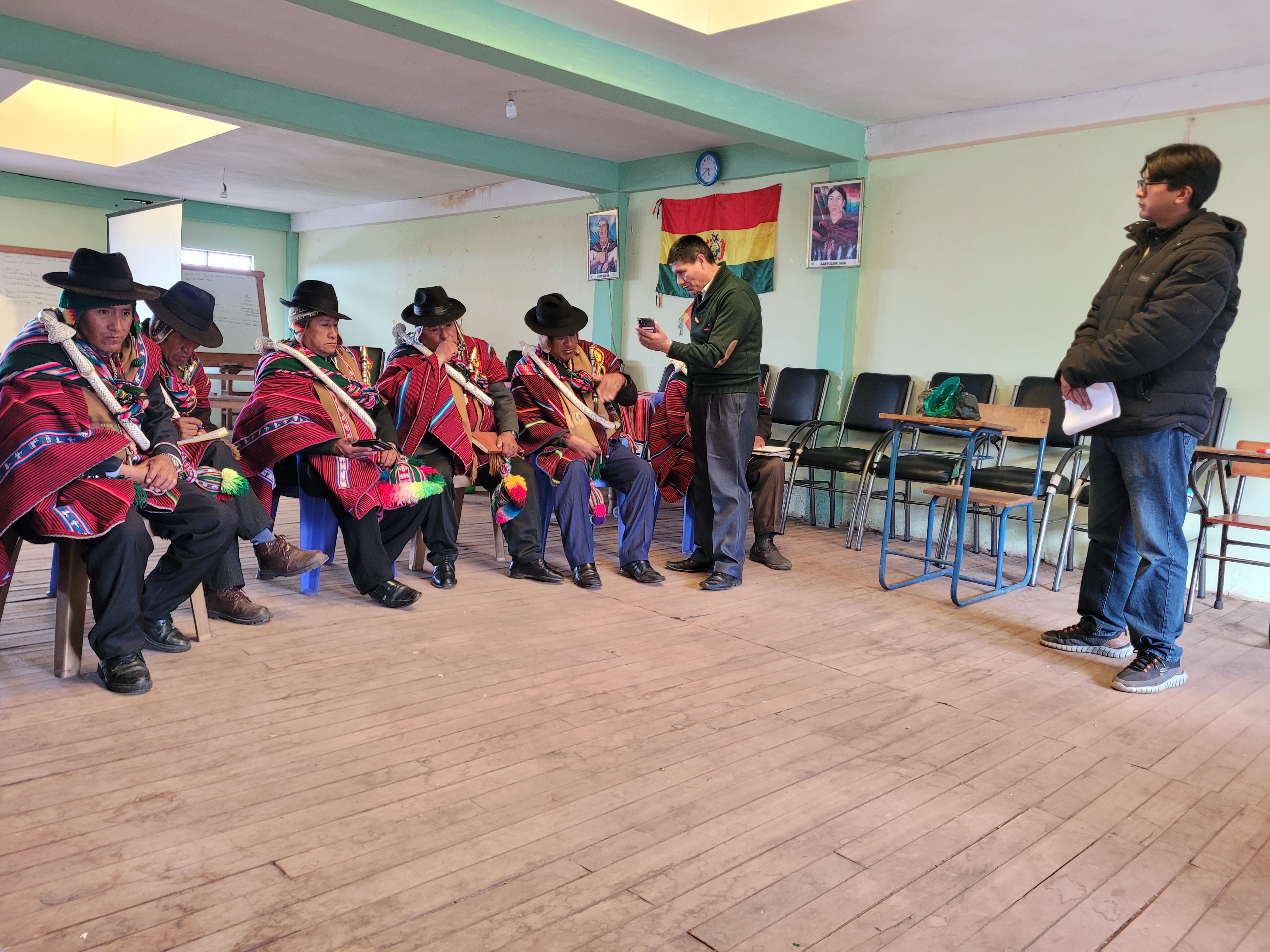 A group of men in traditional Andean clothing sit in a classroom while two men stand and speak, with a Bolivian flag and chairs in the background.