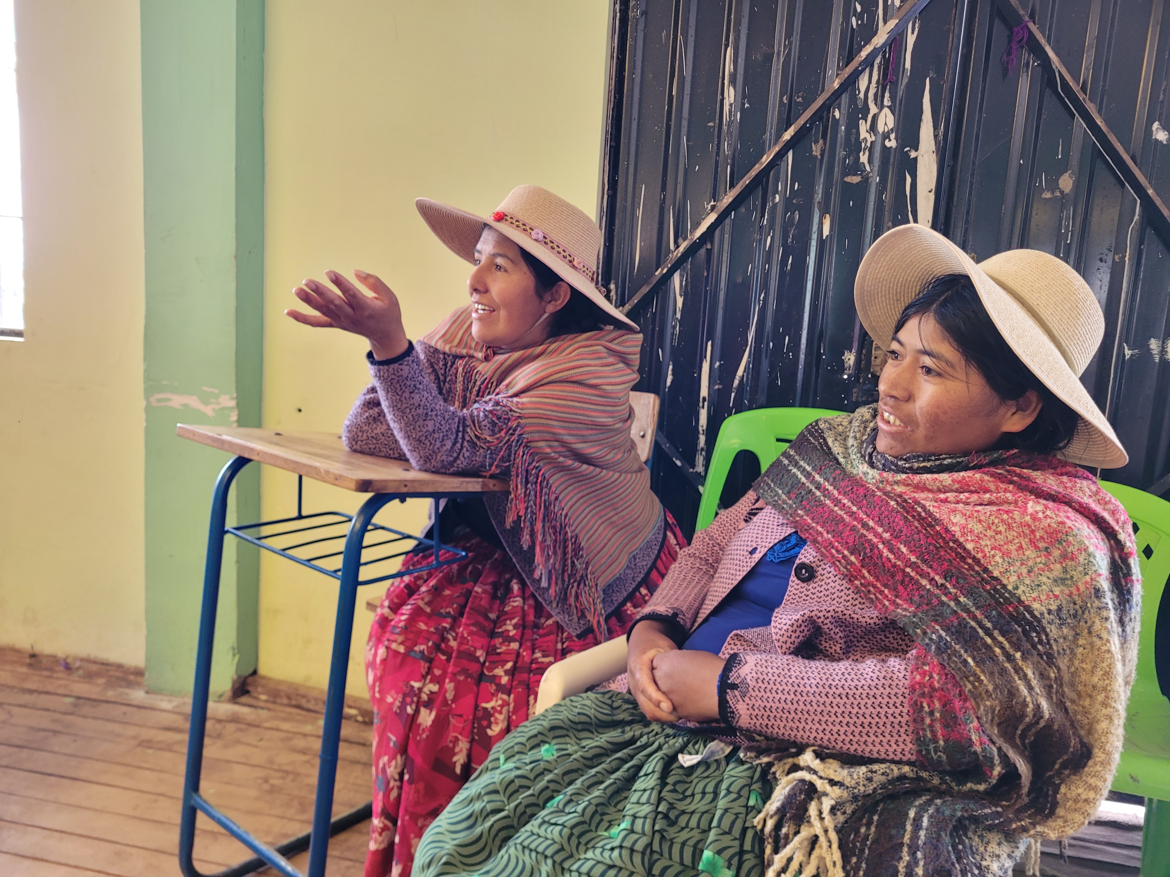 Two women in traditional clothing and hats sit indoors, one at a desk gesturing and smiling, the other sitting next to her, both appearing engaged in conversation.