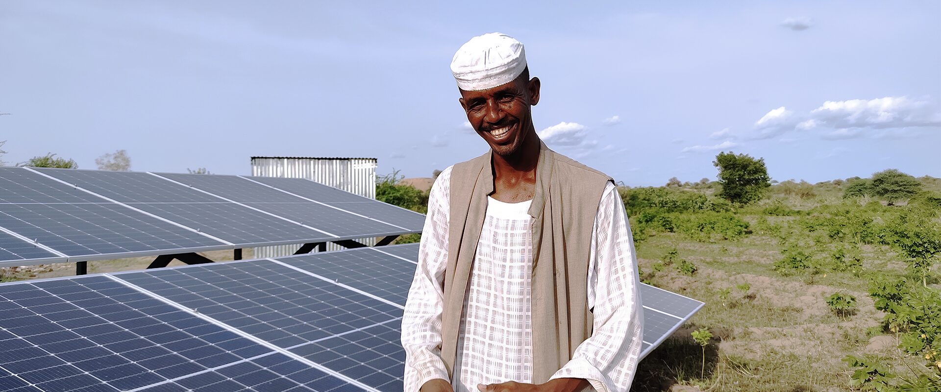 A man stands outdoors smiling in front of several solar panels and greenery under a partly cloudy sky.